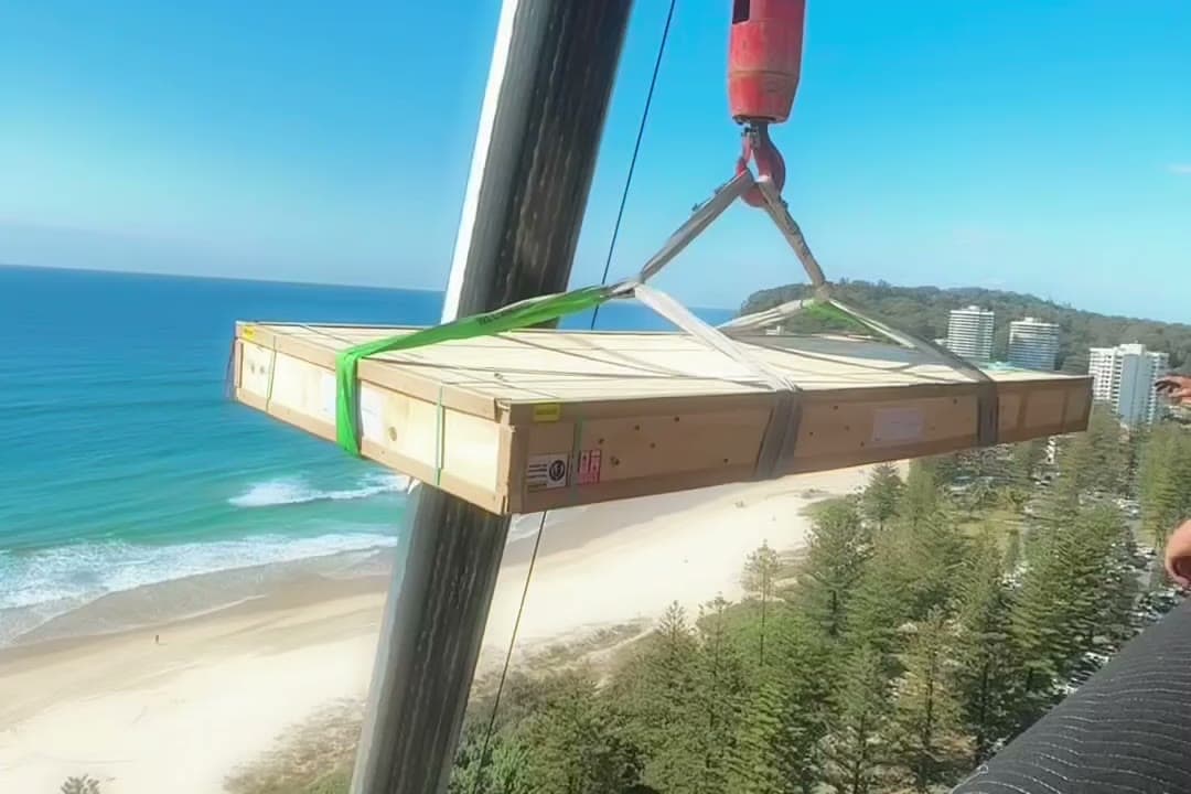 A crated piece lifted by crane to a high-rise balcony above the Gold Coast.