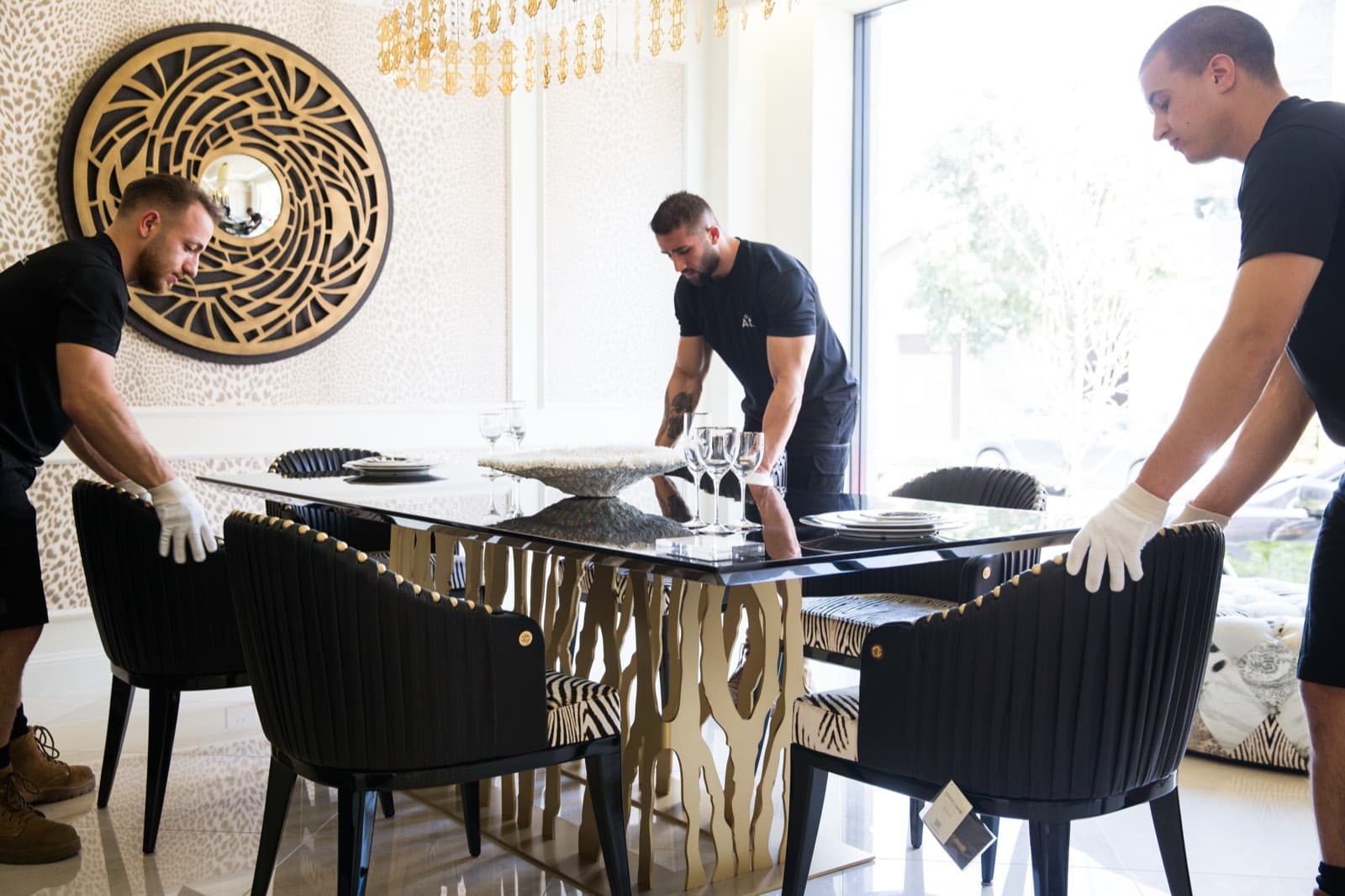 Ateam Logistics handlers setting a sculptural dining table and upholstered chairs into position beneath a gold sunburst mirror.