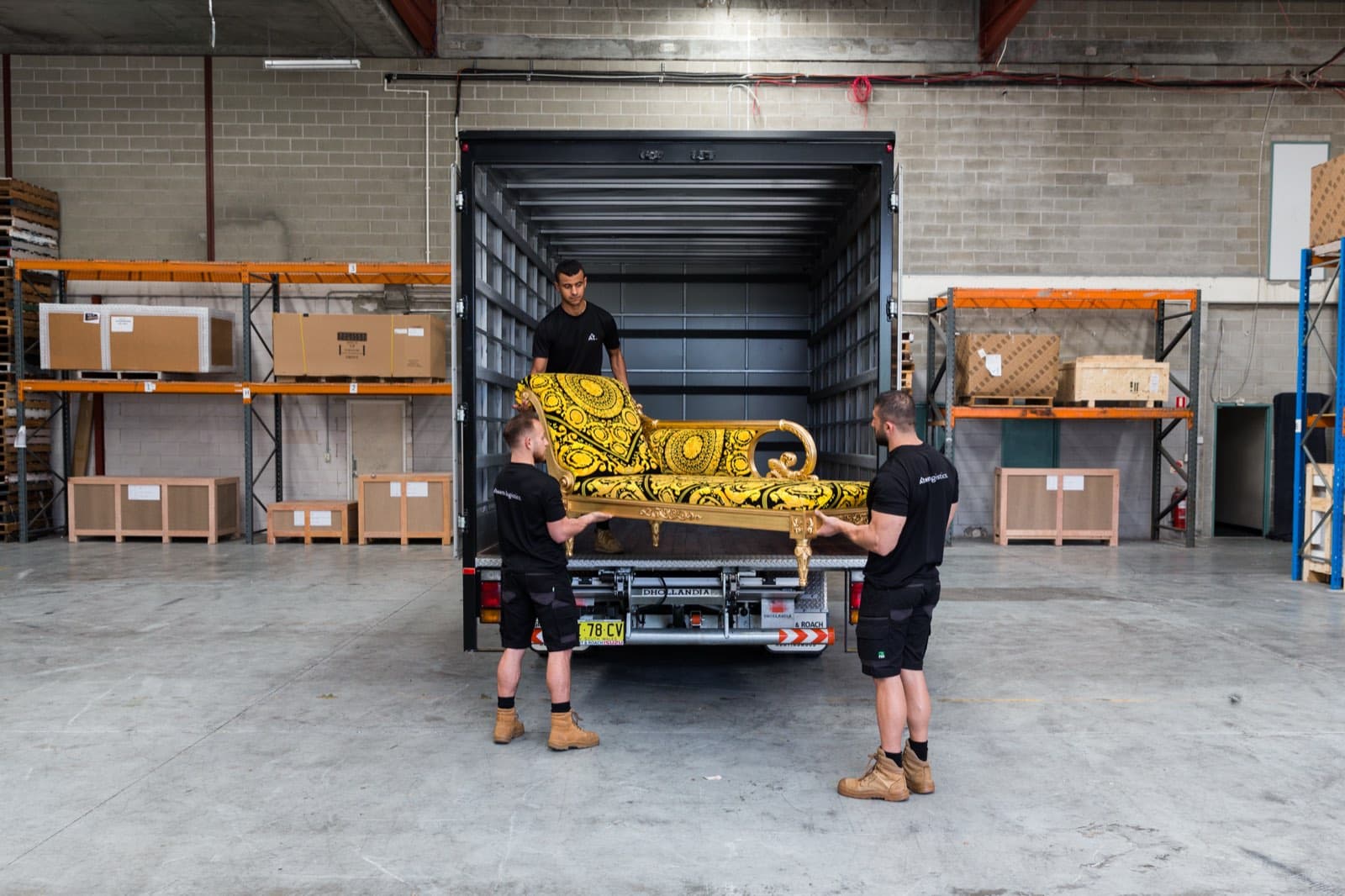 Ateam Logistics handlers lifting an ornate gold-patterned chaise longue onto the tailgate of a matte black Ateam truck at the warehouse loading dock.